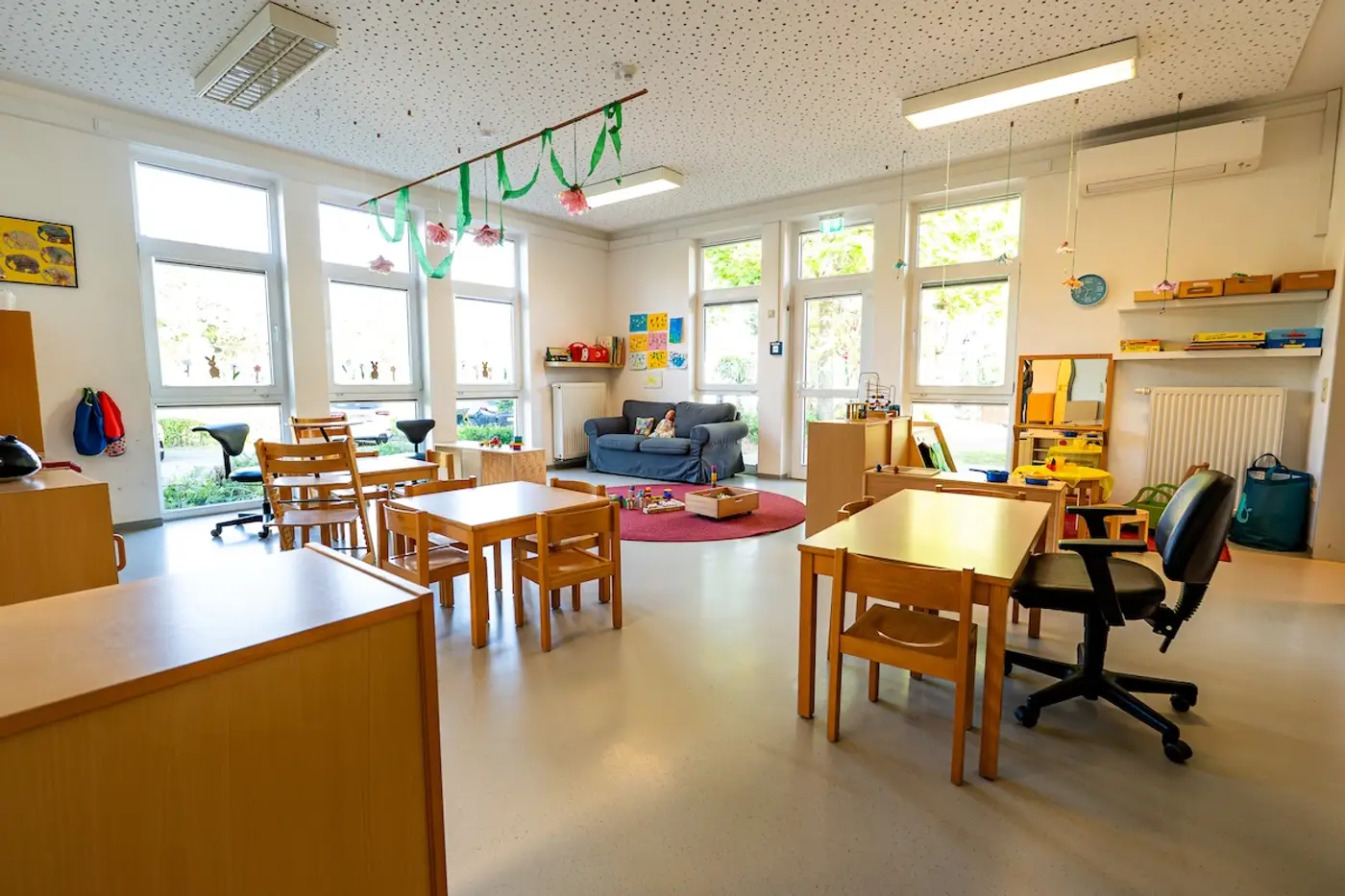 Gemütlicher Gruppenraum der Nestgruppe mit rotem Spielteppich, Sofa und Kindertischen in der Kindertagesstätte Schweich